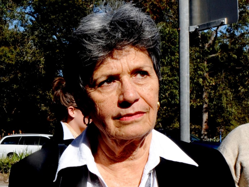 An elderly woman with short black hair, small round earrings, white, shirt and black jacket looks away from the camera. 