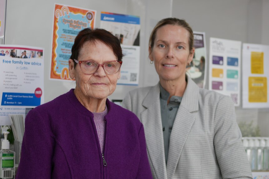 Two women standing, smiling, office background, woman on left is wearing purple and the other is in grey