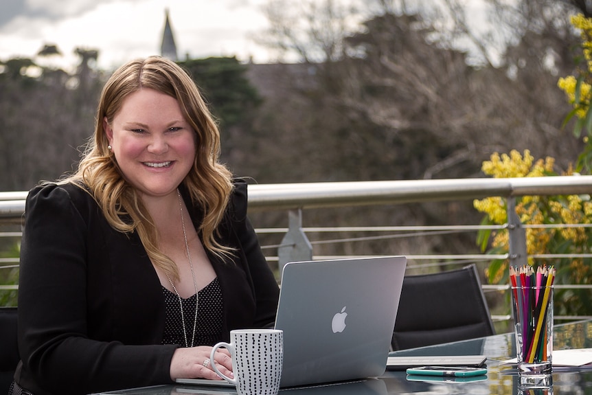 A woman sits at a table outside working on a laptop. She is looking into the lens of the camera and smiling