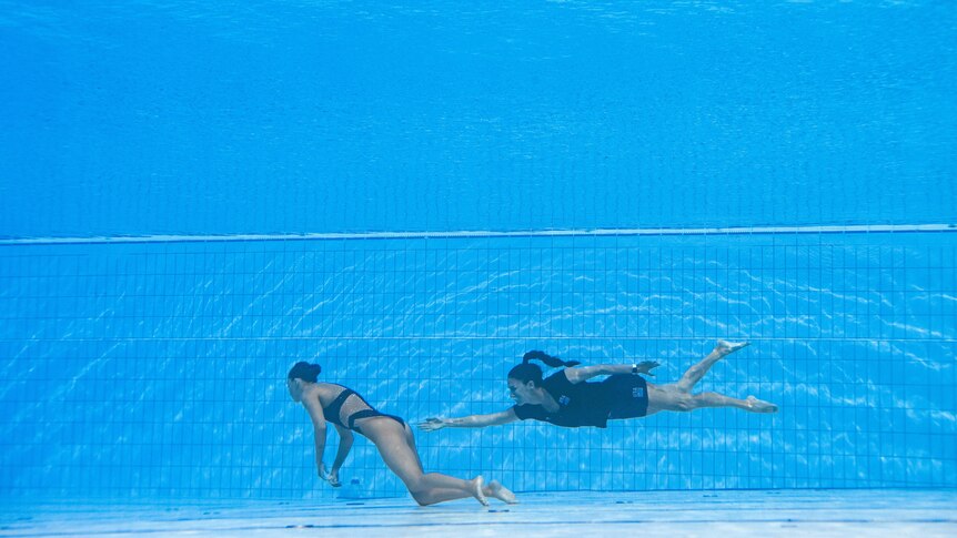 A woman reaches out under water to rescue a swimmer who has fainted at the bottom of a deep pool.