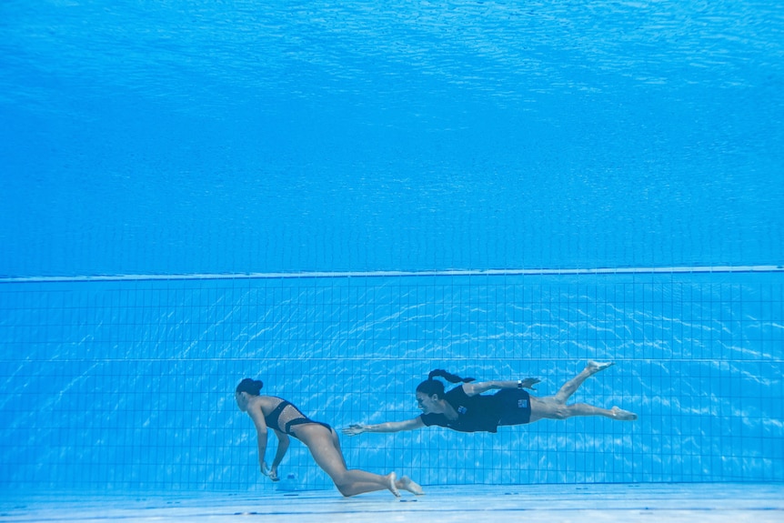 A woman reaches out under water to rescue a swimmer who has fainted at the bottom of a deep pool.