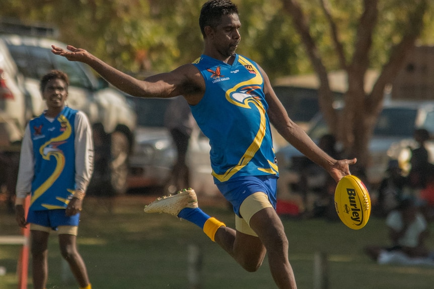 A man in a blue football jumper kicks a yellow Aussie Rules ball, as a teammate watches on