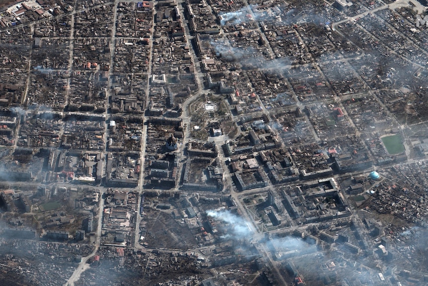 A satellite aerial image of the city of Mariupol with multiple plumes of smoke rising from buildings across the landscape