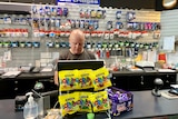 A man behind a busy counter, at a computer, with products lined up on the wall behind him.