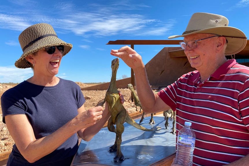 A man and a woman wearing hats laugh while playing with a very small dinosaur statue.