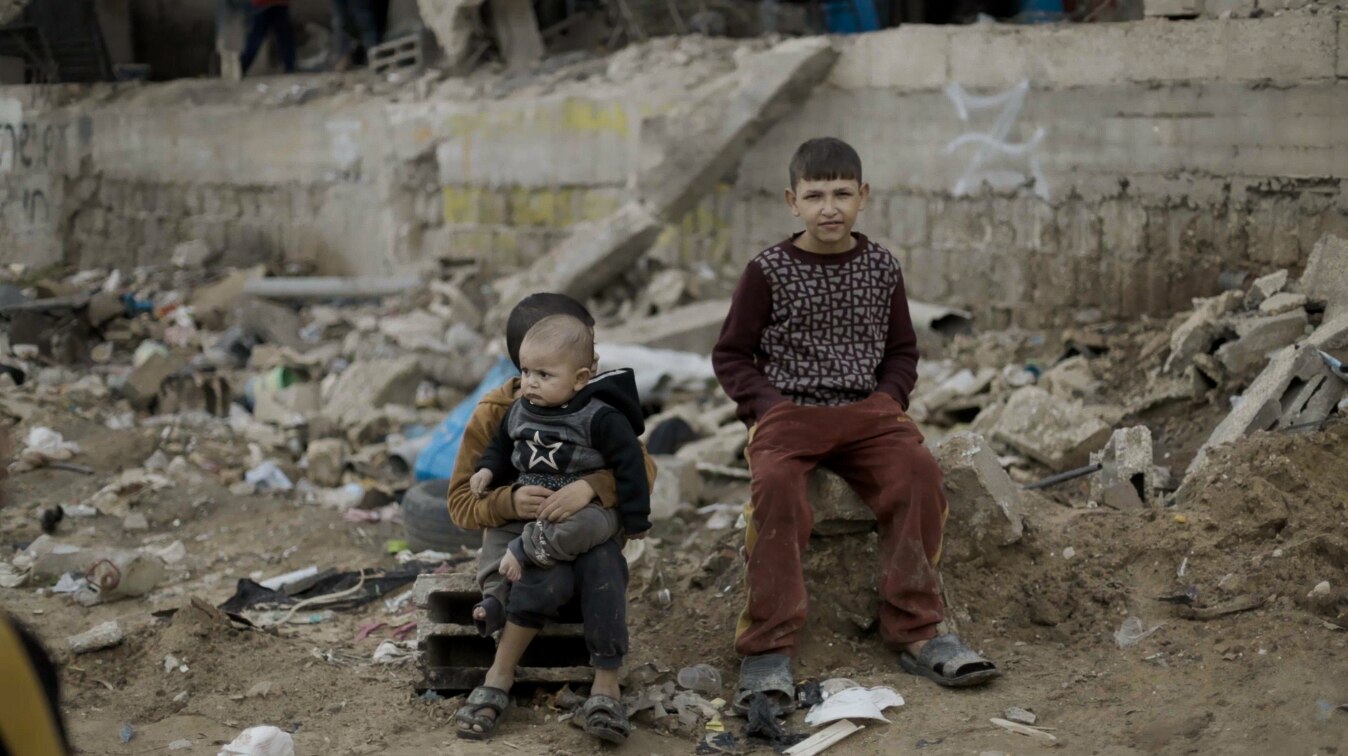 Two boys sit on the ground surrounded by broken bricks, sticks and other debris.