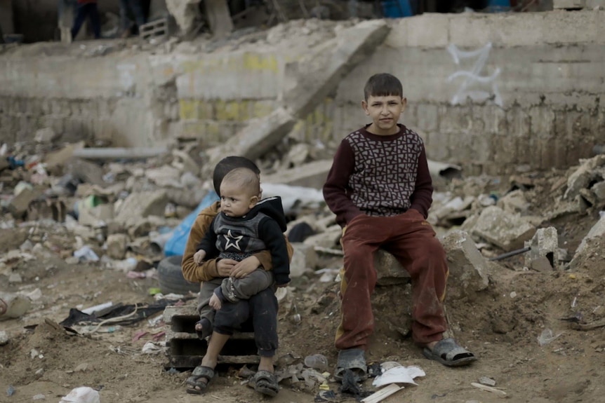 Two boys sit on the ground surrounded by broken bricks, sticks and other debris.