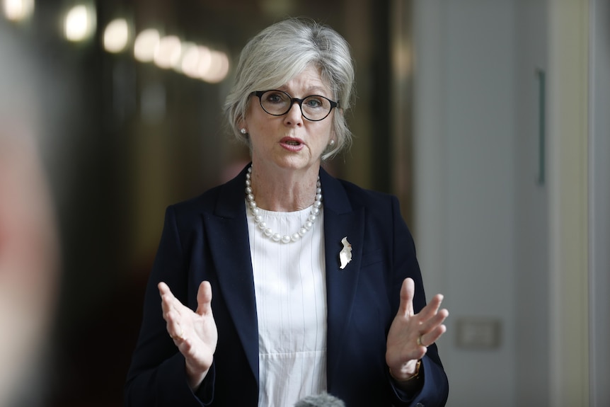 Helen Haines mid-sentence gesturing with her hands open with a string of lights in a corridor out of focus behind her