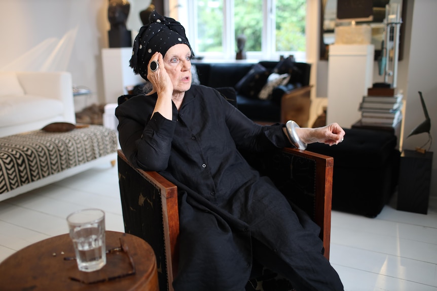 Wendy Whiteley sitting in a chair in a living room with white tiles, she wears black with a black headscarf and large black ring