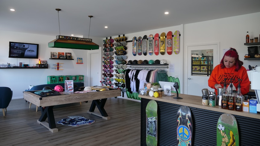 a girl with purple hair wearing an orange jumper is making coffee, there are skateboards on the walls 