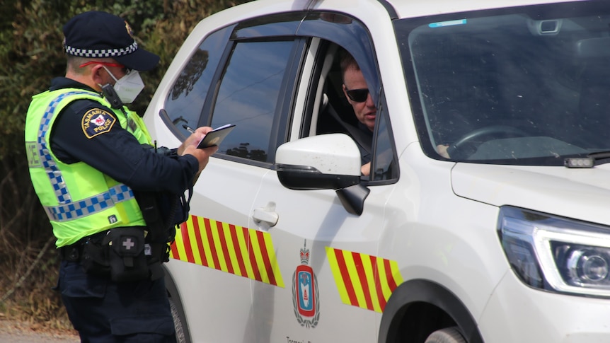 A police officer standing on a country road talks to a fire officer, who is in a car. 