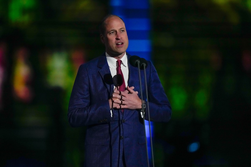 Prince William stands a microphone, both hands on his heart. 