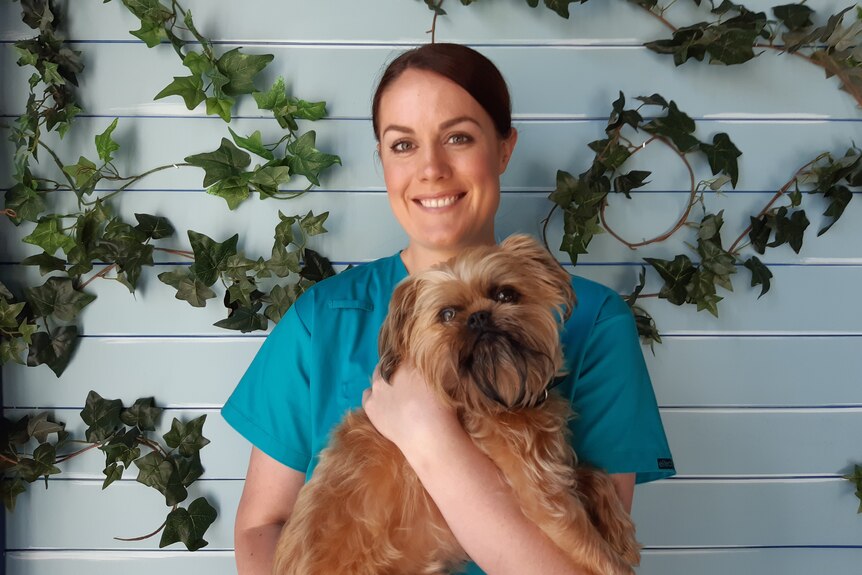 A brunette woman in vet scrubs stands smiling and holding a dog.