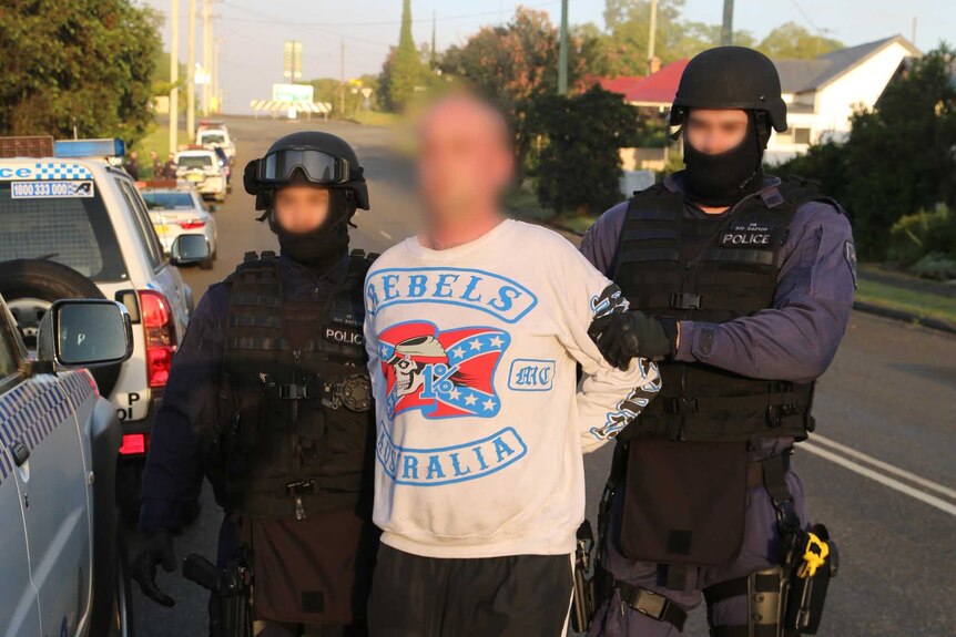 A man in a 'Rebels Australia' jumper is held by two police officers