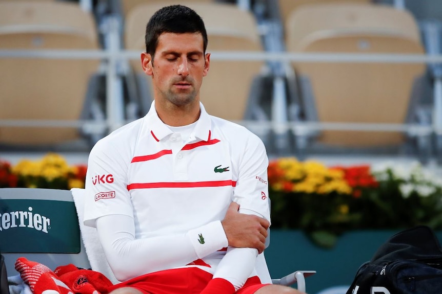 Novak Djokovic clutches at his left arm while sitting in a chair with his eyes closed.