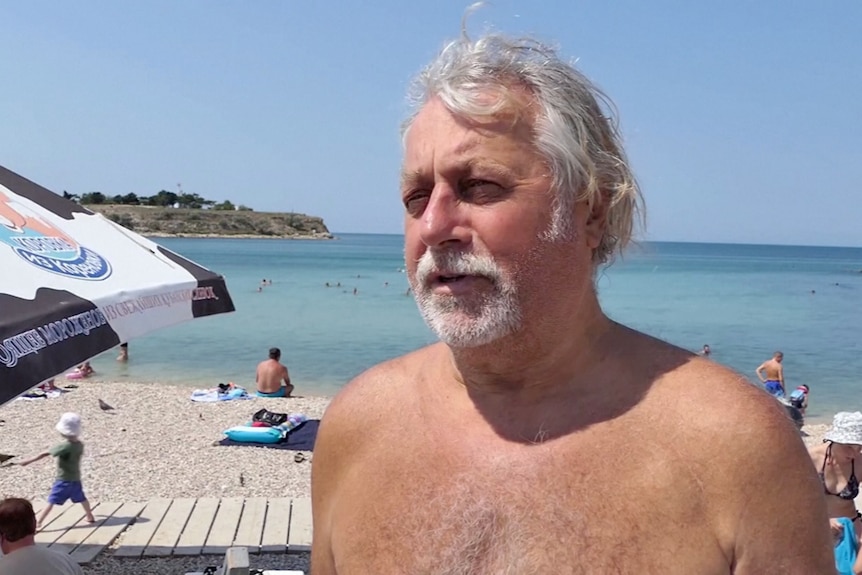 A man with grey hair and a little beard stands shirtless at a beach.