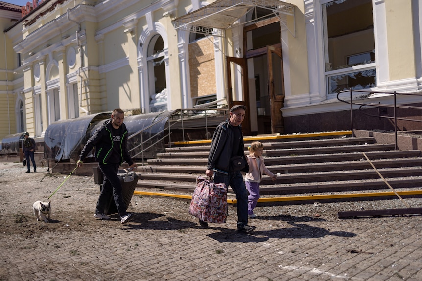 Two men, a young girl and a small dog jog past a ruined entrance to a train station.