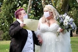 Jessica King in a suit and Emily Sinclair in a white gown wear decorative face masks while colourful confetti falls around them.