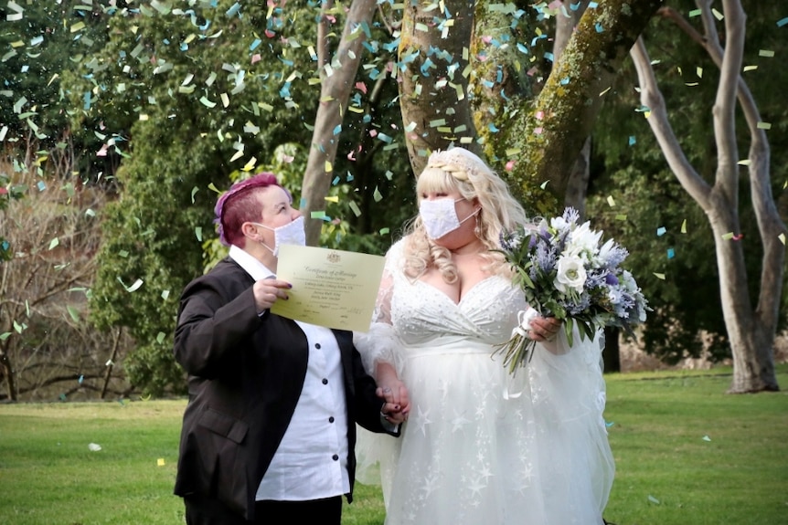 Jessica King in a suit and Emily Sinclair in a white gown wear decorative face masks while colourful confetti falls around them.