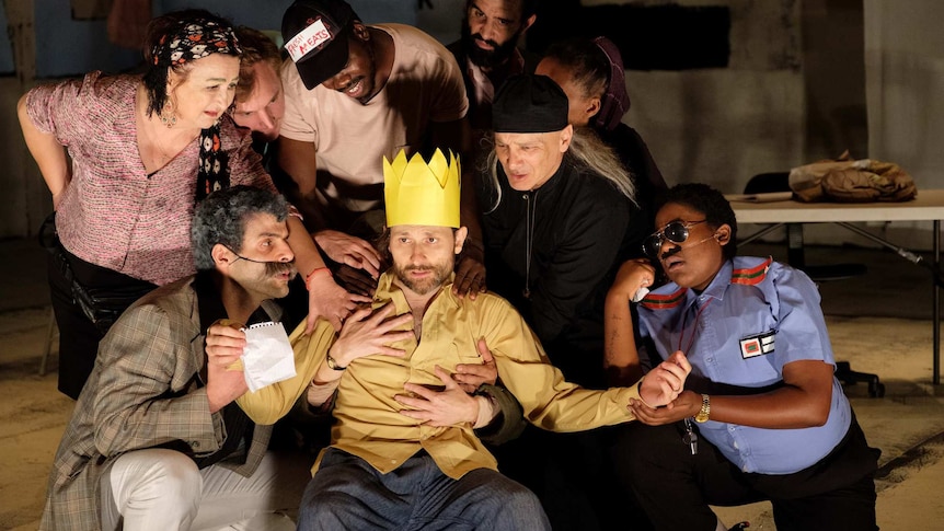 On stage, eight diverse characters gather around Yalin Ozucelik who is wearing a paper crown and looks distraught.