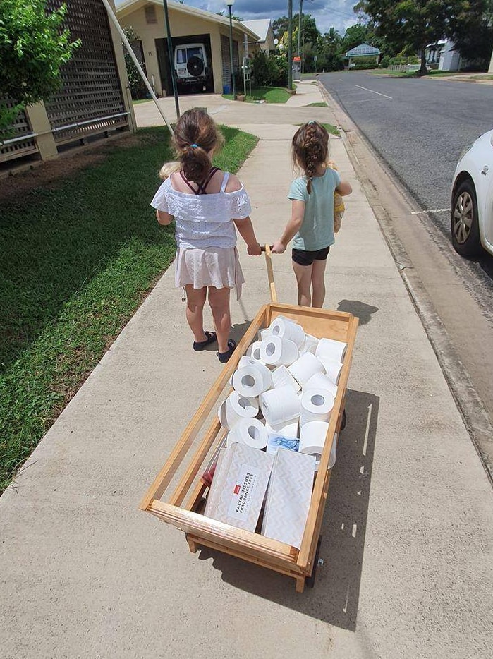 Two little girls pull a wooden wagon with dozens of toilet paper rolls and boxes of tissues.