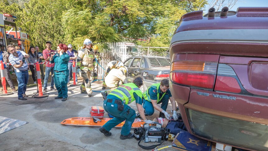 Students stand to the side watching as paramedics remove a dummy from an overturned red car.