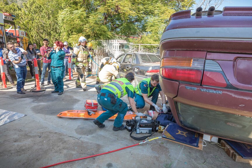 Students stand to the side watching as paramedics remove a dummy from an overturned red car.