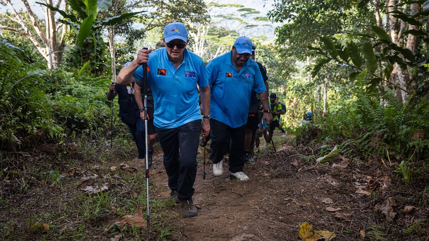 Two men in matching outfits walk closely together along a dirt track, jungle visible on either side.