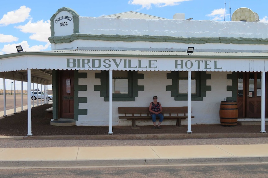 A woman sits on a bench outside a grand old outback pub that reads "Birdsville Hotel - established 1884".