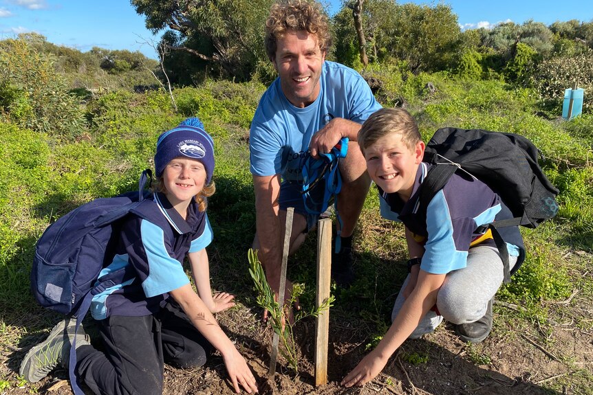 Man in middle crouching down behind seedling planted in ground, flanked by two students, one on left with backpack and beanie 