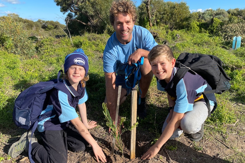 Man in middle crouching down behind seedling planted in ground, flanked by two students, one on left with backpack and beanie 