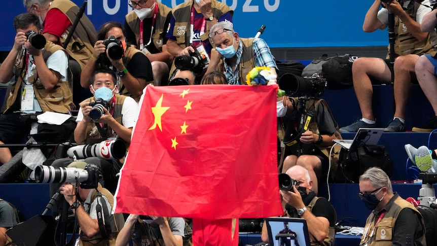 A person unfurls a large Chinese flag at a stadium, many photographers are seen in the background