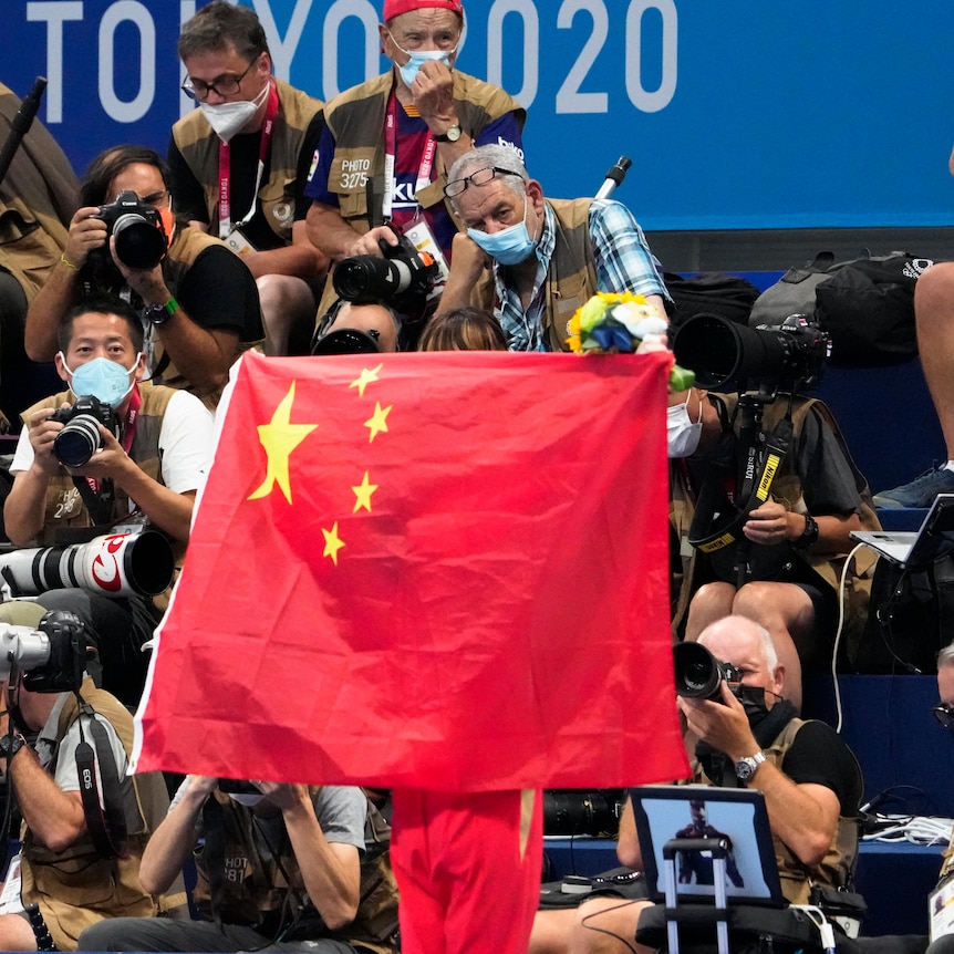 A person unfurls a large Chinese flag at a stadium, many photographers are seen in the background