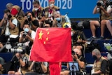 A person unfurls a large Chinese flag at a stadium, many photographers are seen in the background
