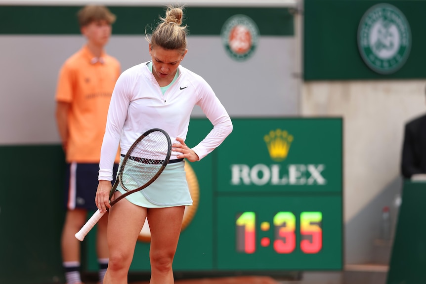 A tennis player wearing pink and green looks down at the court with a racquet in her hand