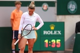 A tennis player wearing pink and green looks down at the court with a racquet in her hand