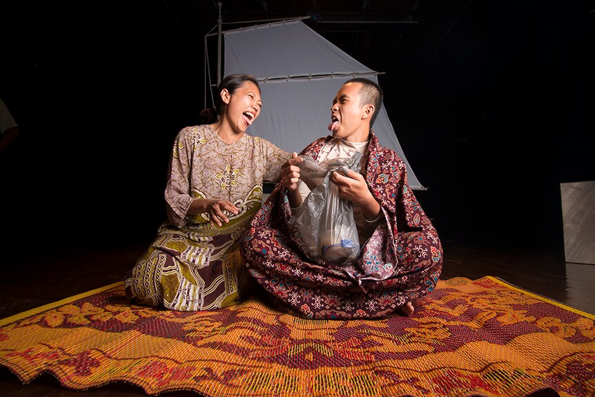 A female actor sitting on a mat laughs with a younger male actor sitting next to her on stage.