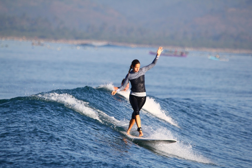 A woman wearing a wetsuit stands on a surfboard, riding a wave.