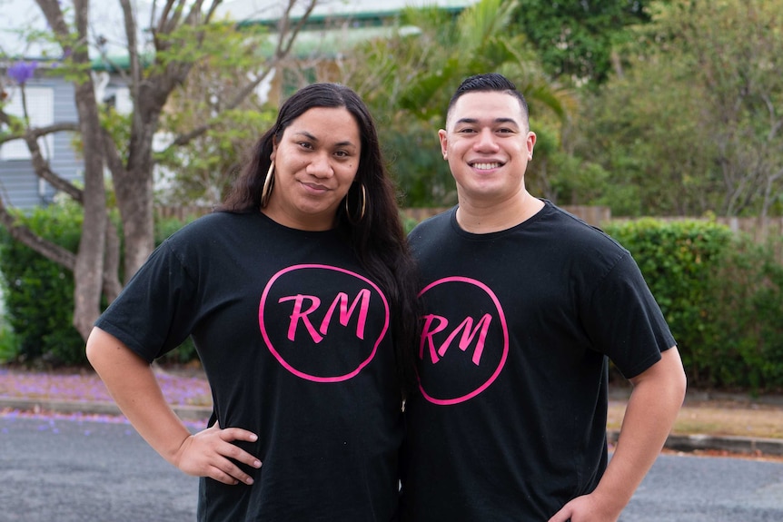 A Samoan transwoman and a young man stand close together on a leafy Brisbane street, smiling.