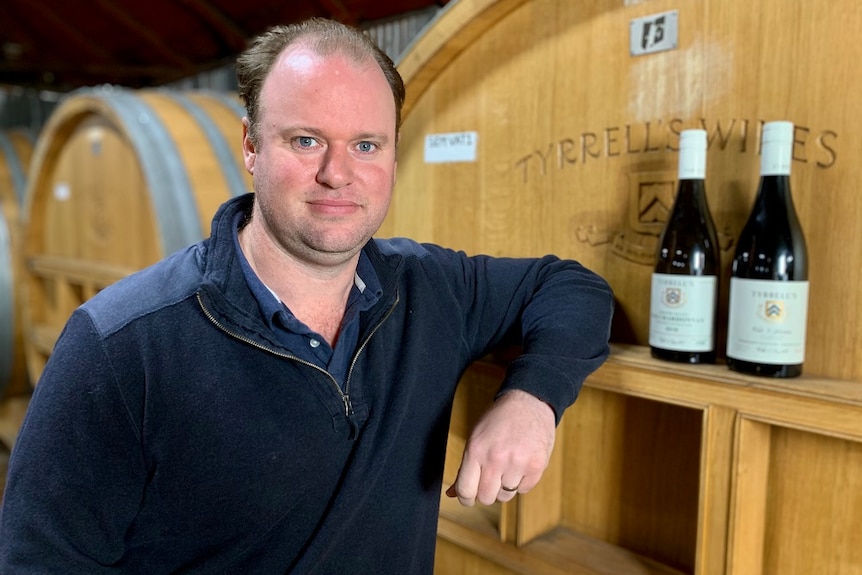 A man standing in the barrel room next to the barrels and two bottles of wine.