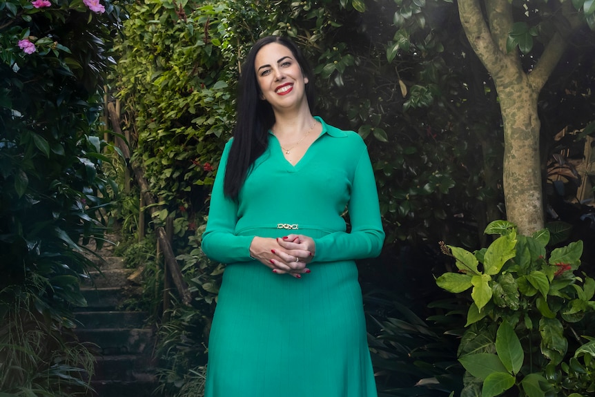 Sarah Holland-Hatt, a 40-year-old woman with long dark hair, stands in nature, smiling. She is wearing a green dress.