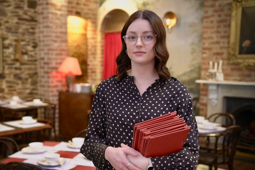 A young Caucasian lady with shoulder length brown hair and glasses stands with menus in her hand inside a restaurant