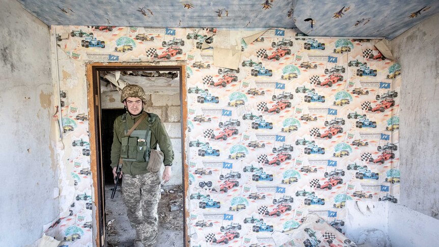 A soldier carrying an automatic rifle walks through a doorway into an abandoned bedroom covered in wallpaper.