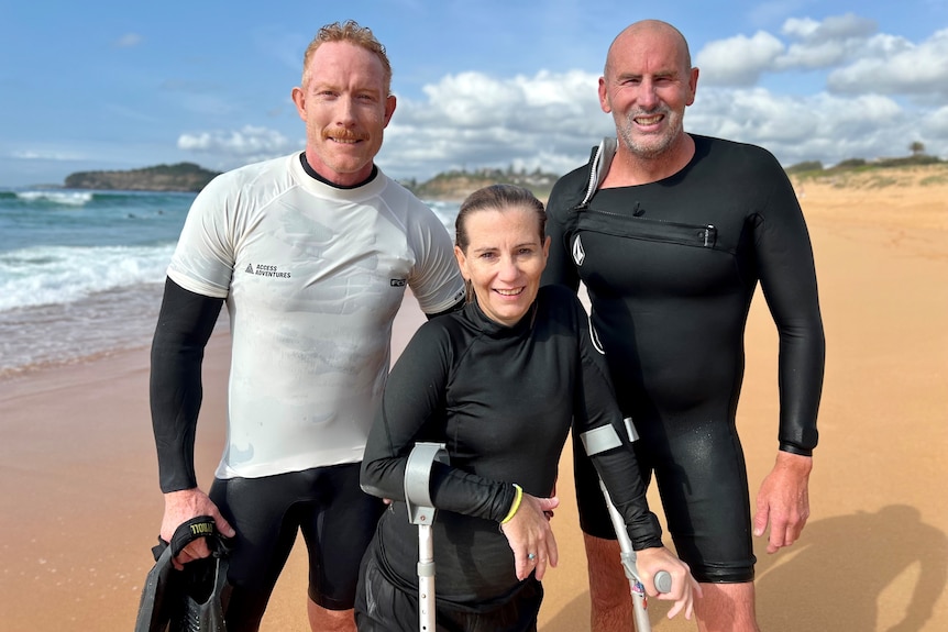 Three people in wetsuits stand on the sand and smile for the camera.