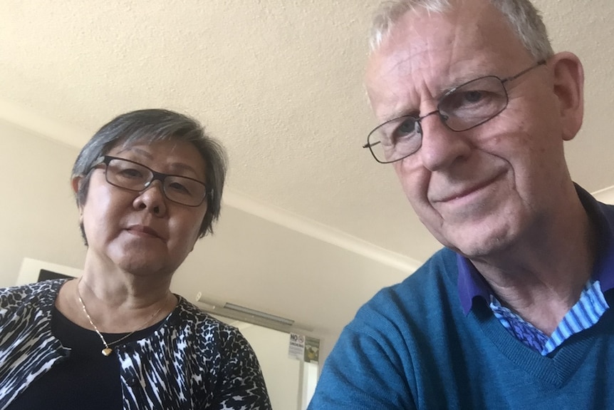 An older man and woman look into the camera from their motel room.