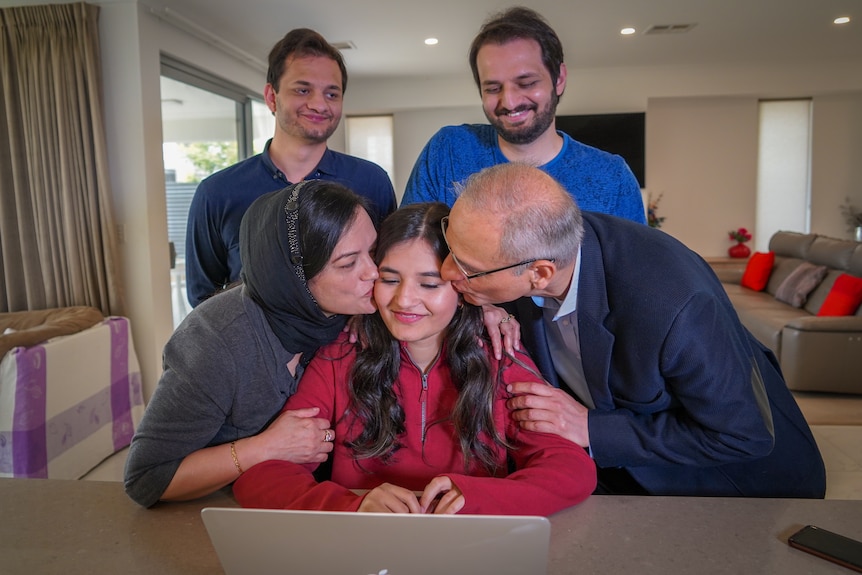 A man and a woman kissing a younger woman looking at a laptop while two other men stand behind