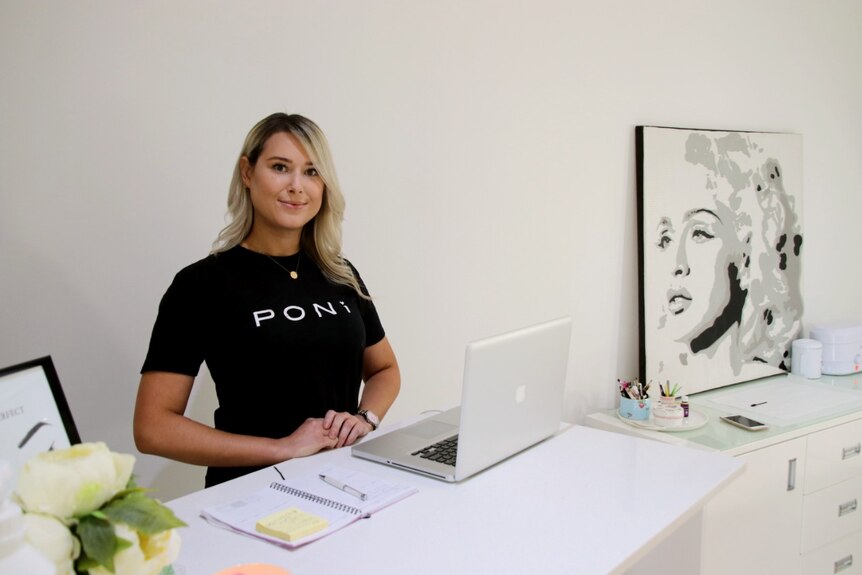 A woman with blonde hair wearing a black t-shirt stands at the counter of a beauty salon with a laptop open in front of her.