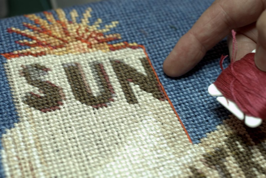 Jessie's finger is pointing close up at her needlepoint work of the Sun Theatre, highlighting the intricacy of her stiches.