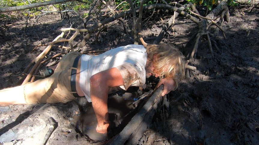 A man lying down in mud with his hand down a hole.