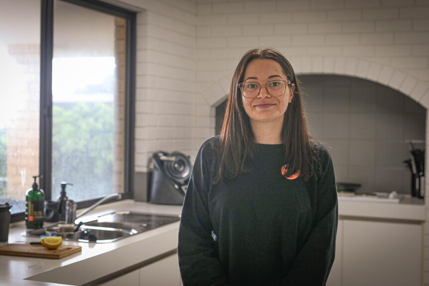 A young woman wearing glasses stands in her kitchen with a small smile to the camera.
